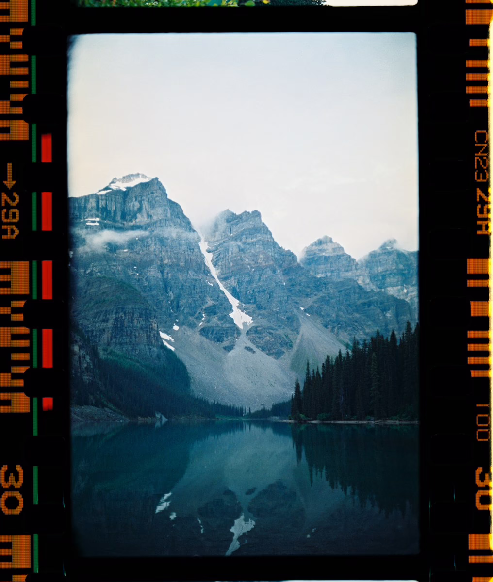 A calm lake reflecting jagged mountains and pine trees under a cloudy sky